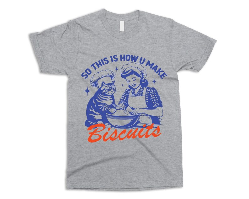 May include: A heather gray t-shirt featuring a blue and orange graphic. The graphic depicts a cat and a woman wearing chef hats, mixing ingredients in a bowl. The text reads "SO THIS IS HOW U MAKE BISCUITS."