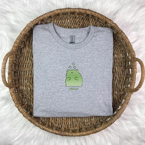 May include: A heather grey t-shirt with a green frog illustration wearing a crown and surrounded by stars. The text "Ribbit" is printed below the frog.