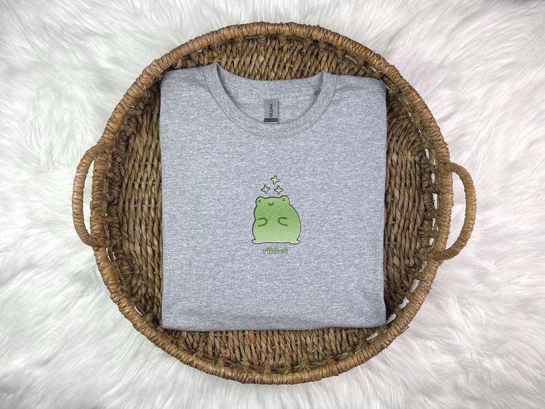 May include: A heather grey t-shirt with a green frog illustration wearing a crown and surrounded by stars. The text "Ribbit" is printed below the frog.