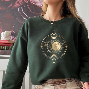 Peut inclure: Un sweat-shirt vert fonc&eacute; avec un motif de lune et de soleil dor&eacute;. Le motif comprend un croissant de lune, une pleine lune et un soleil avec un motif floral.