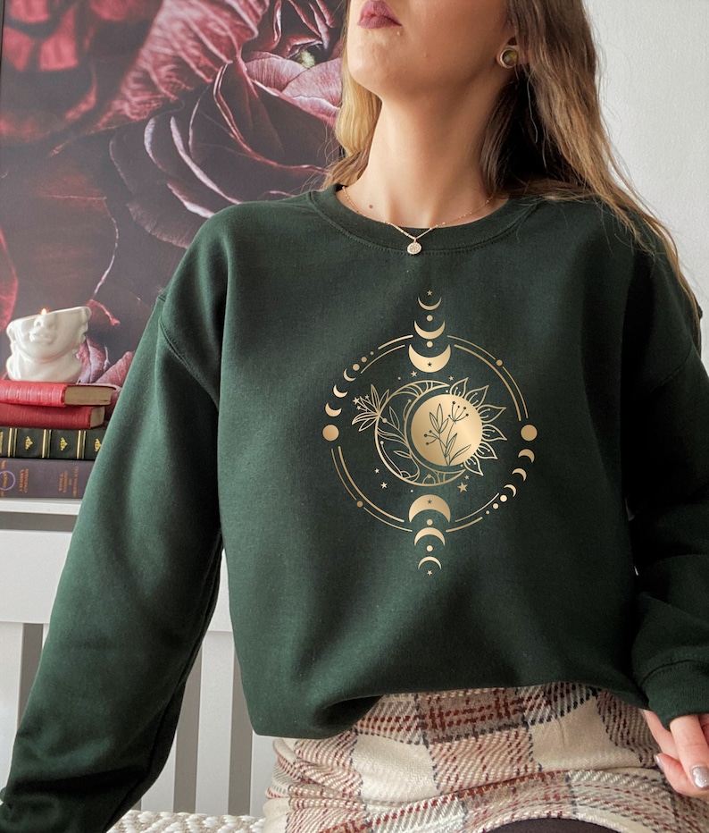 Peut inclure: Un sweat-shirt vert fonc&eacute; avec un motif de lune et de soleil dor&eacute;. Le motif comprend un croissant de lune, une pleine lune et un soleil avec un motif floral.