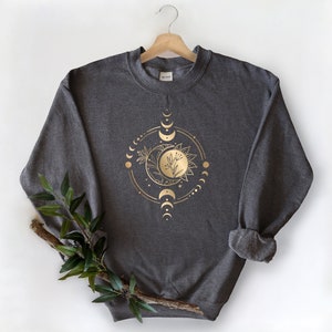Peut inclure: Un sweat-shirt gris fonc&eacute; avec un motif en feuille d'or d'une lune, d'un soleil et d'&eacute;l&eacute;ments floraux dans un cercle.