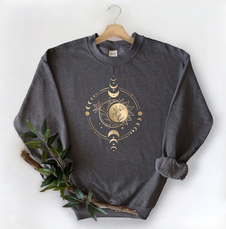 Peut inclure: Un sweat-shirt gris fonc&eacute; avec un motif en feuille d'or d'une lune, d'un soleil et d'&eacute;l&eacute;ments floraux dans un cercle.