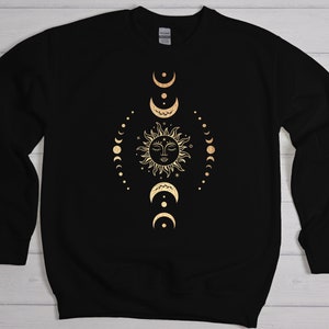 May include: Black sweatshirt with a gold sun and moon design. The sun has a face and is surrounded by a circle of small stars. The moon phases are arranged in a vertical line around the sun.