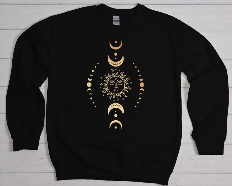 May include: Black sweatshirt with a gold sun and moon design. The sun has a face and is surrounded by a circle of small stars. The moon phases are arranged in a vertical line around the sun.