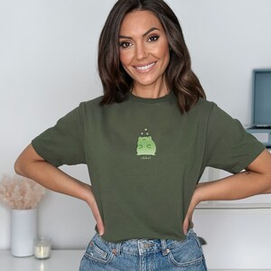 May include: A woman wearing a green t-shirt with a green frog graphic and the text "ribbit!" printed on it. She is smiling and looking at the camera.