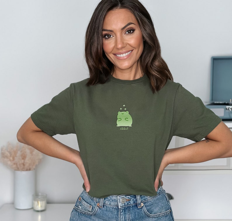 May include: A woman wearing a green t-shirt with a green frog graphic and the text "ribbit!" printed on it. She is smiling and looking at the camera.