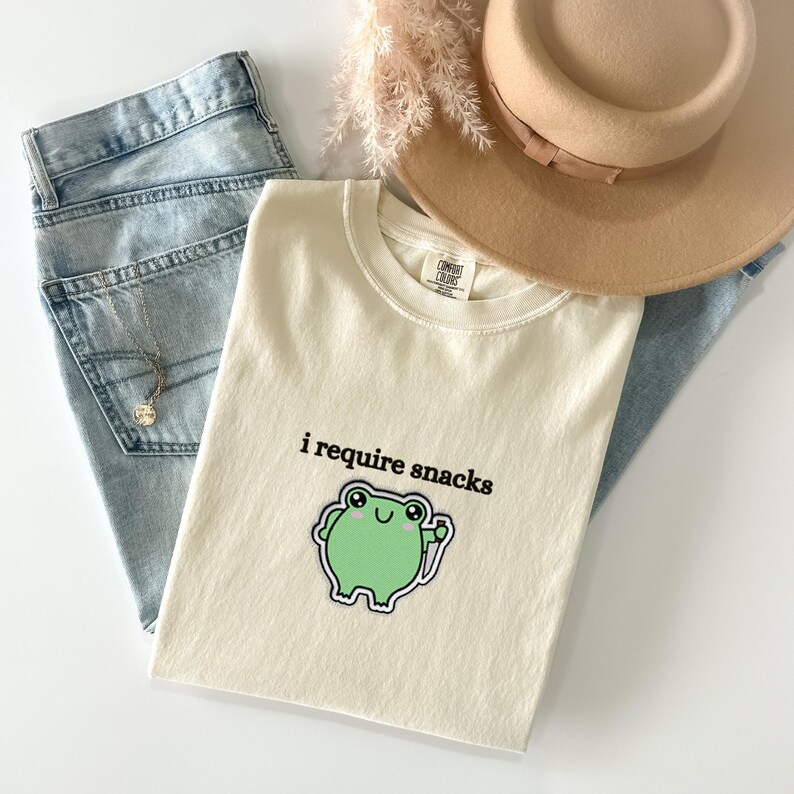 May include: Cream-colored t-shirt with the phrase "i require snacks" and a green frog graphic. The shirt is paired with light wash denim jeans, a tan hat, and a necklace. A casual, everyday outfit.