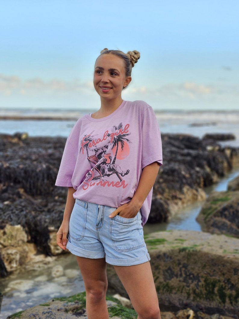 May include: A woman wearing a light pink t-shirt with the text "Feral Girl Summer" in pink and black. She is wearing light blue denim shorts and has her hair pulled back in two buns.