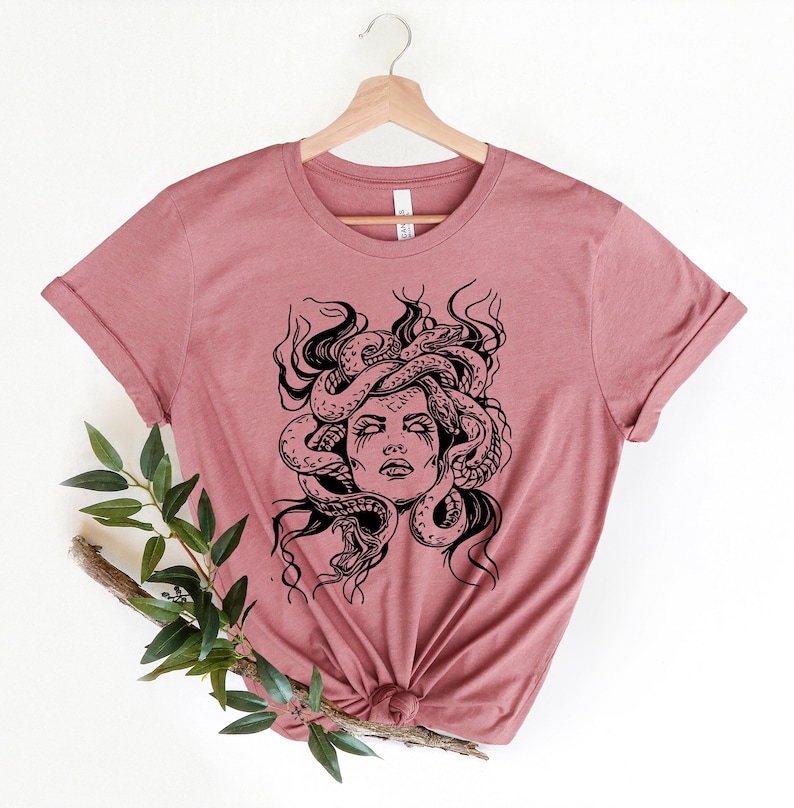 May include: A mauve t-shirt with a black and white graphic print of Medusa, a mythical Greek figure with snakes for hair.