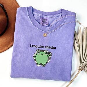 May include: A lavender Comfort Colors t-shirt with the phrase "i require snacks" and a green frog graphic. A gold necklace and a brown hat are also in the image.