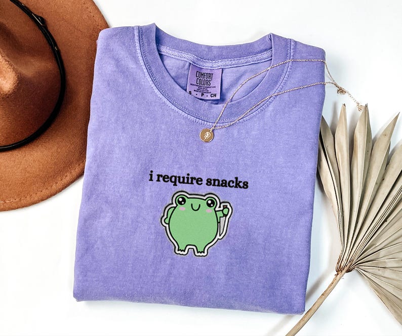 May include: A lavender Comfort Colors t-shirt with the phrase "i require snacks" and a green frog graphic. A gold necklace and a brown hat are also in the image.