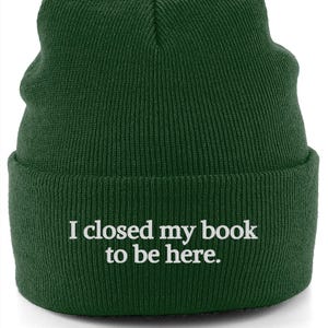 May include: A dark green beanie with the embroidered text "I closed my book to be here." The hat is made of a knitted material and has a folded brim. The text is in a white, sans-serif font.
