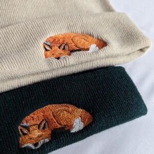 May include: Two knit beanies with a fox design embroidered on the front. The top beanie is beige and the bottom beanie is dark green.