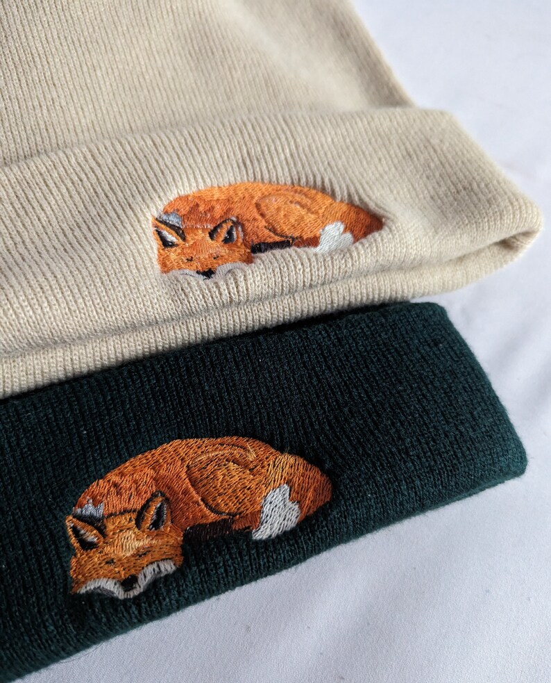 May include: Two knit beanies with a fox design embroidered on the front. The top beanie is beige and the bottom beanie is dark green.
