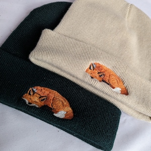 May include: Two knit beanies with fox embroidery. One beanie is beige and the other is dark green. Both foxes are orange and white.
