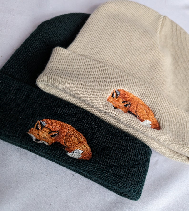 May include: Two knit beanies with fox embroidery. One beanie is beige and the other is dark green. Both foxes are orange and white.