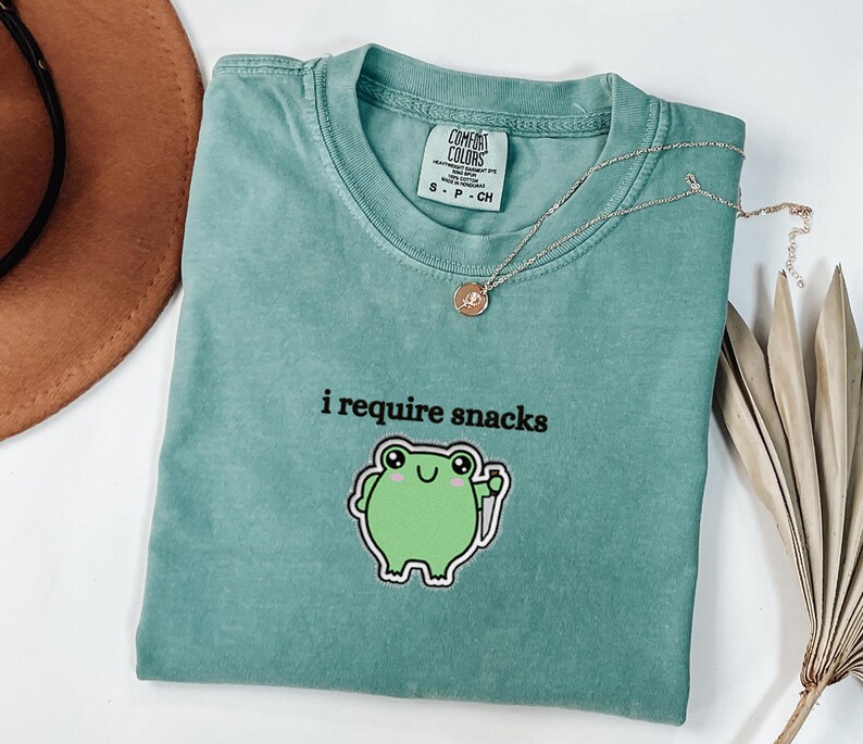 May include: A teal Comfort Colors t-shirt featuring the text "i require snacks" and a green frog illustration. A silver necklace with a pendant is on the shirt. A brown hat and dried plant are in the background.