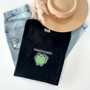 May include: A black t-shirt with the text "I require snacks" and a green frog graphic. The shirt is laid on top of light blue jeans and a tan hat. A gold necklace is visible on the jeans.