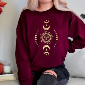 May include: A maroon sweatshirt with a gold sun and moon design. The sun has a face and is surrounded by a circle of small moons.