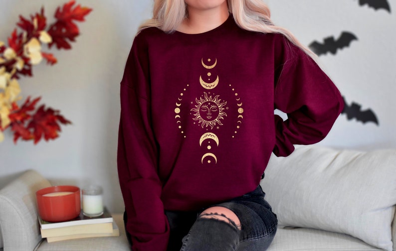 May include: A maroon sweatshirt with a gold sun and moon design. The sun has a face and is surrounded by a circle of small moons.