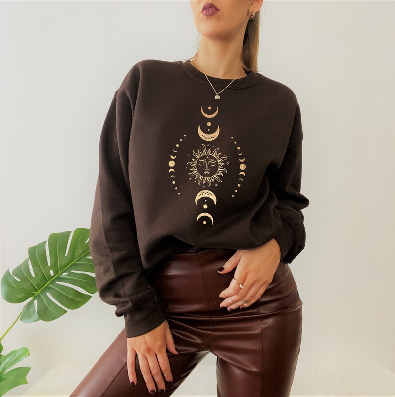 May include: A brown sweatshirt with a gold sun and moon design. The design features a sun with a face and a series of crescent moons.