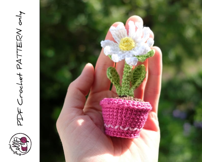 May include: A crocheted daisy flower in a pink pot. The flower is white with a yellow center and green leaves. The pot is crocheted with a pink yarn and has a white trim.