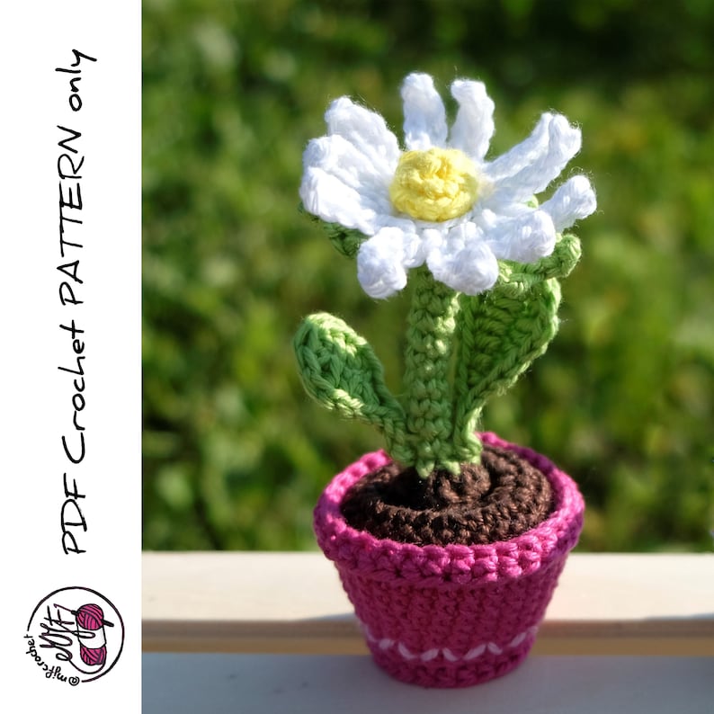 May include: A crocheted daisy flower in a pink pot. The flower is white with a yellow center and green leaves. The pot is a bright pink with a brown crocheted base.