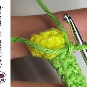 May include: A close-up of a crochet project with a yellow flower and green leaves. The crochet hook is visible in the image.