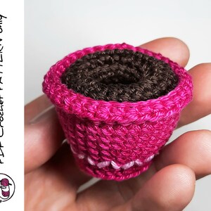 May include: A small pink crocheted flower pot with a brown crocheted center. The pot is being held in a hand.