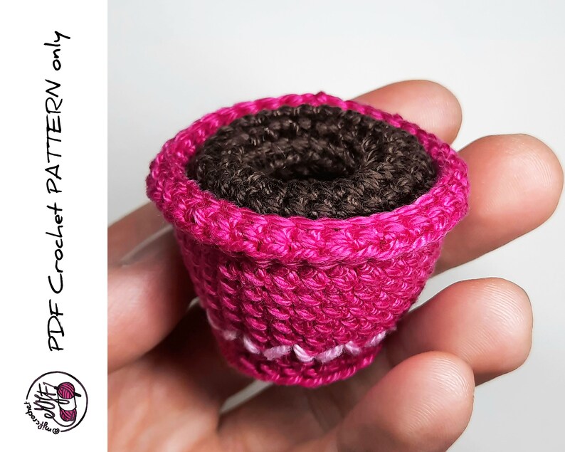 May include: A small pink crocheted flower pot with a brown crocheted center. The pot is being held in a hand.