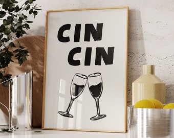 May include: A framed print with the text "Cin Cin" in black lettering on a white background. Two black silhouettes of wine glasses clinking together are below the text. The print is on a white countertop with a clear glass pitcher and a silver bowl with lemons.