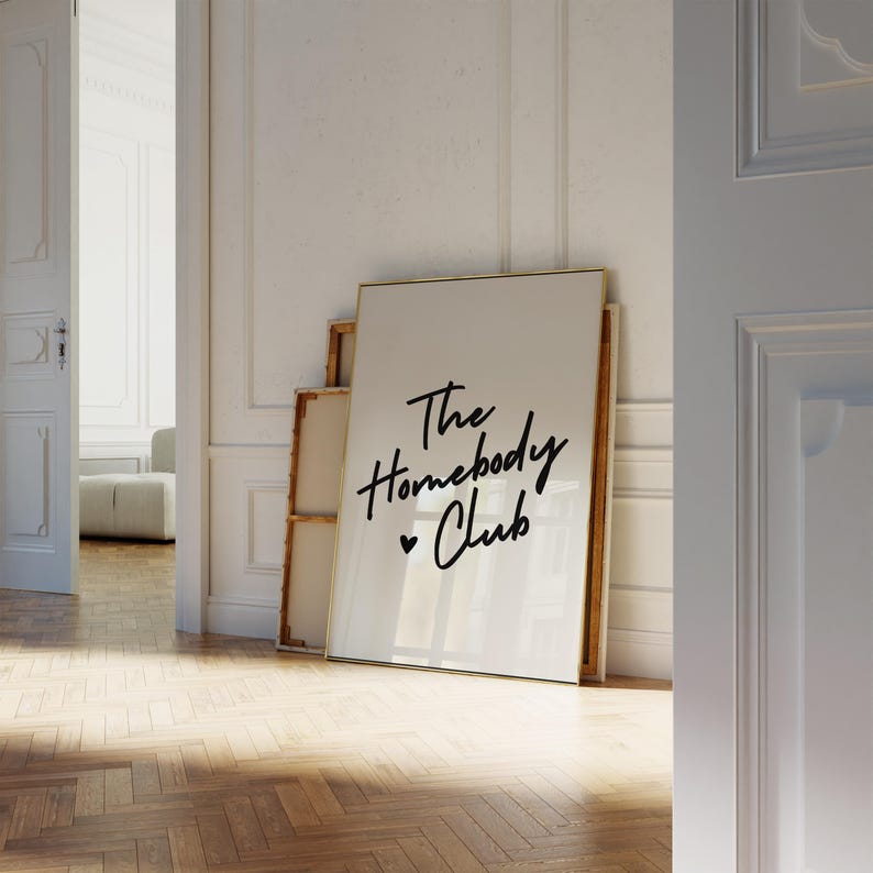 May include: A white framed poster with the text "The Homebody Club" in black cursive lettering with a small heart symbol.