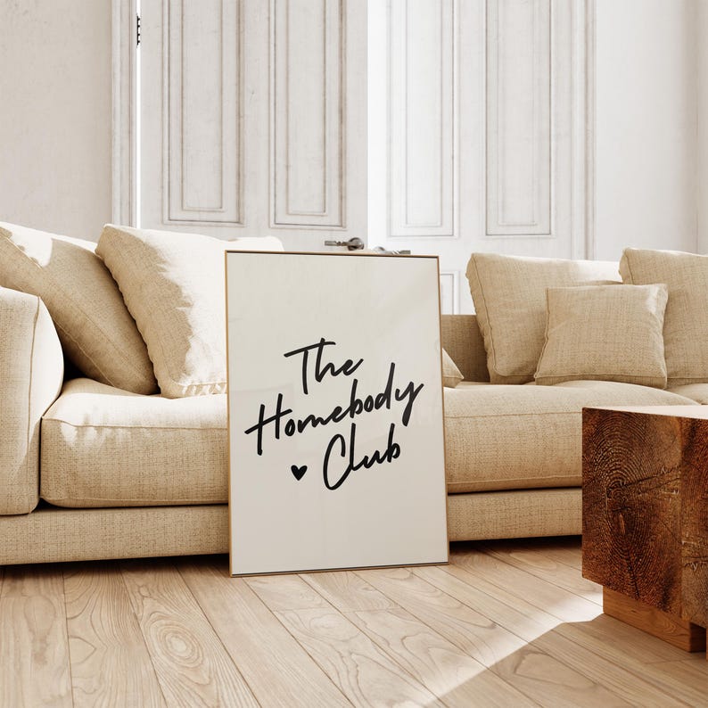 May include: A white poster with a gold frame, featuring black text that reads "The Homebody Club" with a small heart symbol.