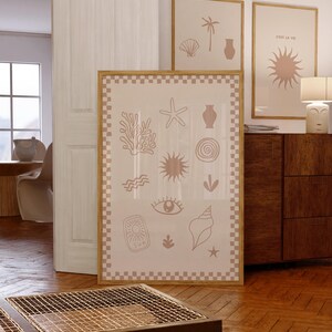 May include: A beige framed print with a checkered border features a collection of hand-drawn illustrations of beach and ocean-themed elements, including a starfish, seashell, sun, eye, and coral. The print is hanging on a white wall in a room with a wooden floor.