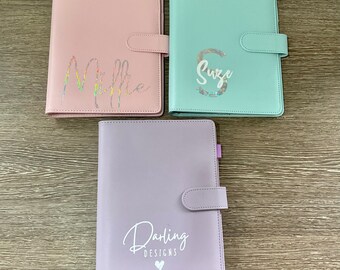 May include: Three pastel-colored leather-bound journals with a snap closure. The journals are pink, light blue, and lavender. The pink journal has the name "Millie" written in iridescent glitter. The light blue journal has the name "Suze" written in iridescent glitter. The lavender journal has the text "Darling Designs" written in white with a small white heart.