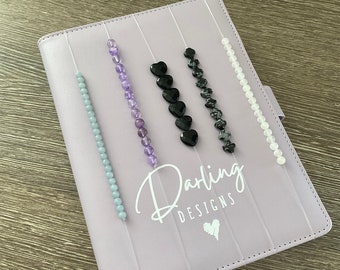May include: A purple notebook with five rows of beads and black heart charms. The notebook has the text "Darling Designs" printed on the front.
