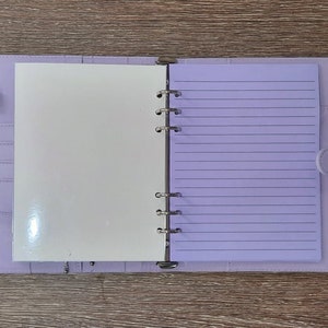 May include: A lavender-colored binder with a white page on the left and a lined page on the right. The binder has a metal ring mechanism and a flap closure.
