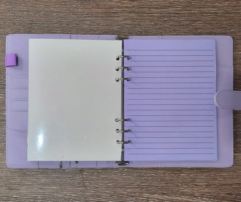 May include: A lavender-colored binder with a white page on the left and a lined page on the right. The binder has a metal ring mechanism and a flap closure.