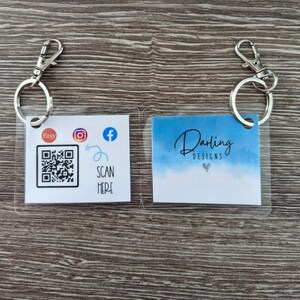 May include: Two keychains with clear plastic holders. The left keychain has a QR code, the text "Etsy", and icons for Instagram and Facebook. The text "SCAN HERE" is below the QR code. The right keychain has a blue watercolour background with the text "Darling Designs".