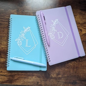 May include: Two spiral bound notebooks with a floral design and a letter in a geometric frame. The notebooks are light blue and lavender. A white pen is on top of the light blue notebook.