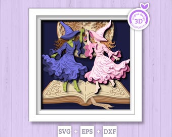 May include: A 3D paper art piece featuring two witches dancing above an open book. One witch wears a purple dress and hat, the other a pink dress and hat. The background is dark blue with a moon, and the artwork is framed in white.