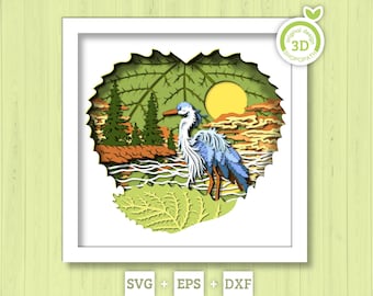 Puede incluir: Una obra de arte en papel en capas que representa una garza azul cerca del agua, con árboles y un sol amarillo en el fondo. La obra está enmarcada en un cuadrado blanco, con un recorte en forma de hoja. Las palabras "SVG + EPS + DXF" están en la parte inferior.