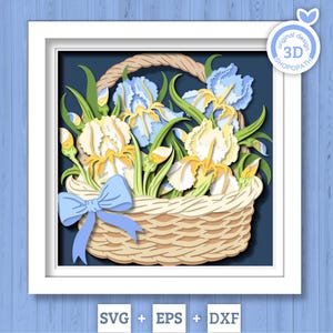 Puede incluir: Una obra de arte en papel 3D de una cesta rebosante de lirios. Las flores son blancas y azules, con hojas y capullos verdes. Un lazo azul claro adorna la cesta. La obra está enmarcada en blanco sobre un fondo azul veteado.