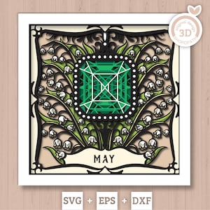 May include: A layered paper art piece featuring a green gemstone at the center, surrounded by lily of the valley flowers. The design includes the word "MAY" and is framed with intricate black and beige details. The bottom corner displays "SVG + EPS + DXF".