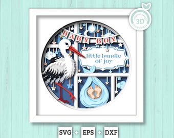 Op de afbeelding: Een gelaagde papieren schaduwdoos met een witte ooievaar met rode snavel en poten, een blauw babypakketje met zichtbare voetjes en banners met de tekst BABY BOY en little bundle of joy tegen een donkerblauwe achtergrond met sterren.