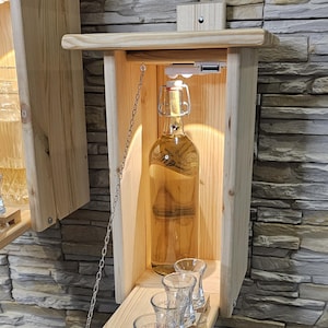 Könnte beinhalten: Ein wandmontierter Likörschrank aus Holz mit einer Flügeltür, die sich öffnet und eine Flasche Likör und ein Licht freigibt. Der Schrank hat eine ausziehbare Ablage, auf der vier Schnapsgläser Platz finden.