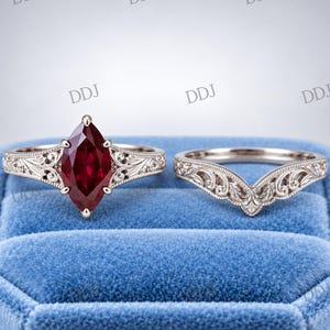 Può includere: Un set di due anelli con una gemma rosso scuro taglio marquise incastonata in una fascia color argento con intricate incisioni a volute, accompagnato da una fede sagomata a forma di V con simili dettagliati motivi in metallo.