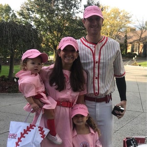 Puede incluir: Una familia de cuatro personas vestida con uniformes de b&eacute;isbol rosa y blanco para Halloween. Los adultos llevan gorras de b&eacute;isbol rosas con el texto "Team DeFeo" en ellas. Los ni&ntilde;os llevan gorras de b&eacute;isbol rosas con el texto "Team DeFeo" en ellas. La familia est&aacute; de pie en una acera frente a una casa.