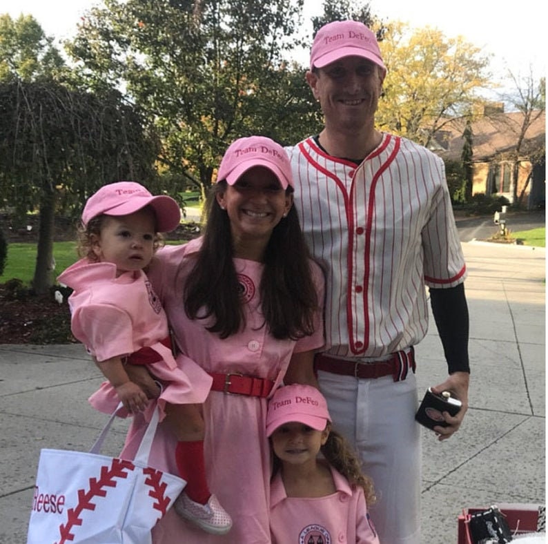 Puede incluir: Una familia de cuatro personas vestida con uniformes de b&eacute;isbol rosa y blanco para Halloween. Los adultos llevan gorras de b&eacute;isbol rosas con el texto "Team DeFeo" en ellas. Los ni&ntilde;os llevan gorras de b&eacute;isbol rosas con el texto "Team DeFeo" en ellas. La familia est&aacute; de pie en una acera frente a una casa.